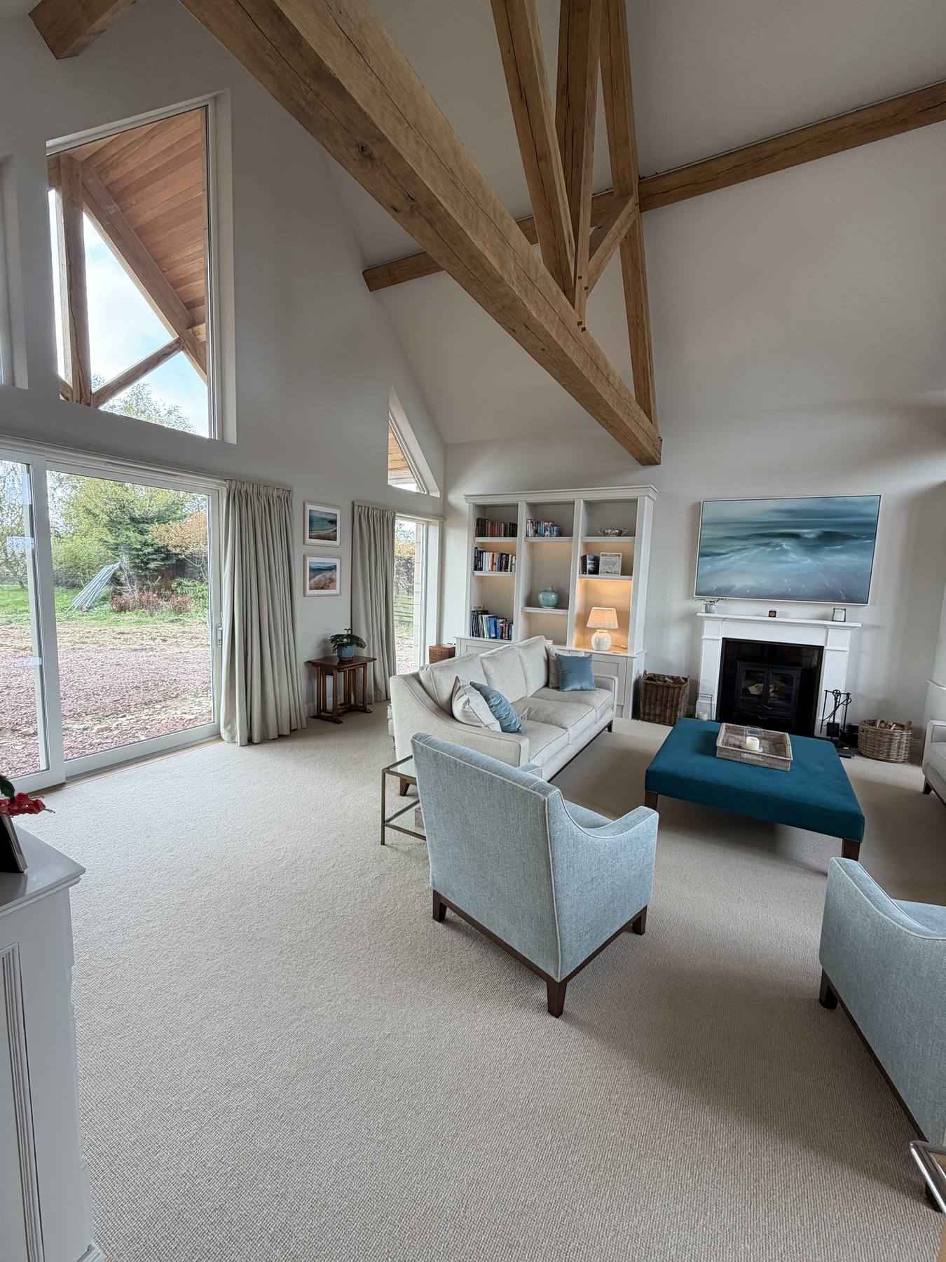 Sitting room with oak trusses, full height glazing, linen curtains, a teal footstool and white shelving beside the fireplace