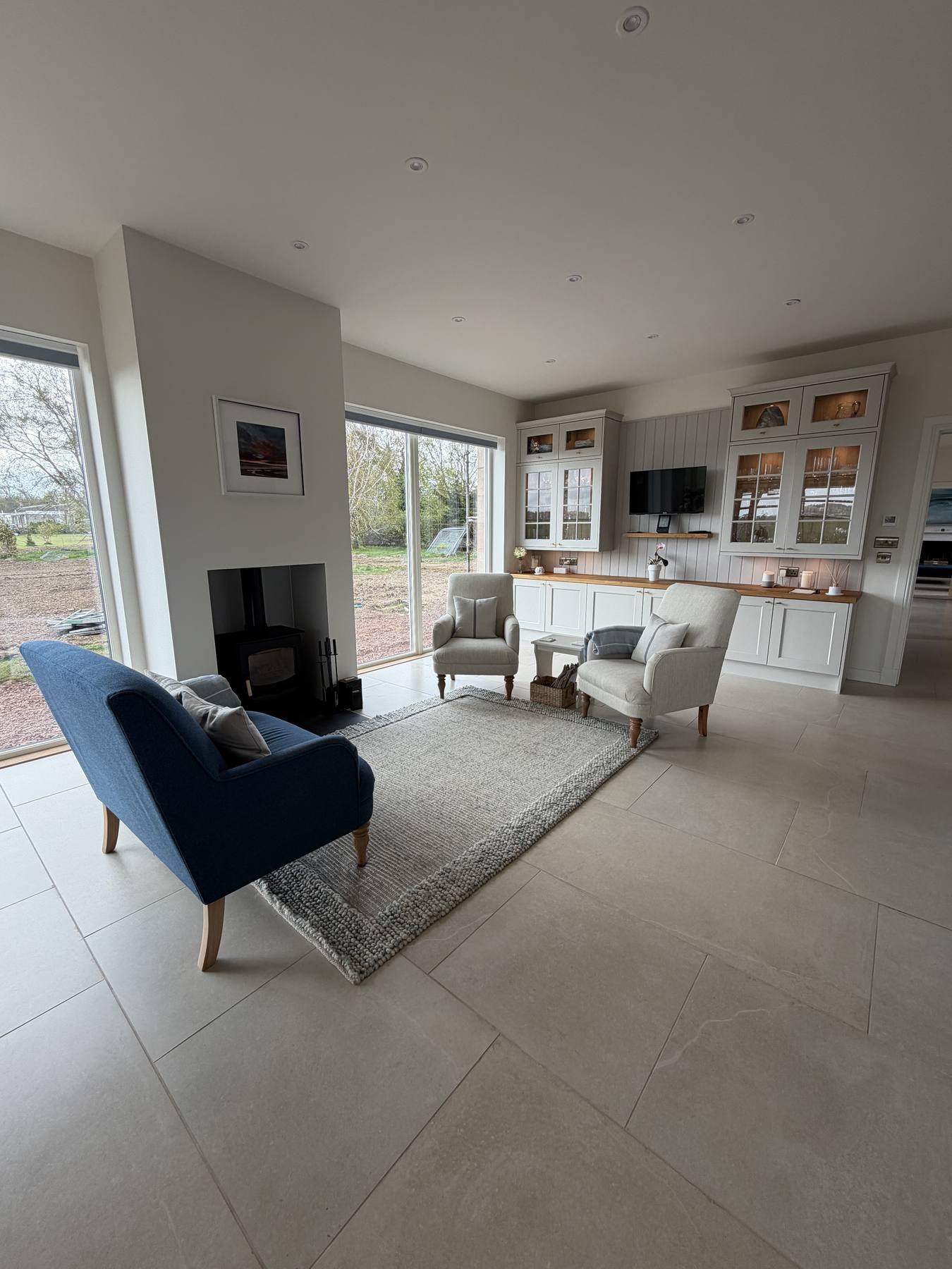 Open plan family room with stone floor tiles, a wood burning stove, a blue armchair, a grey rug and white kitchen cabinetry